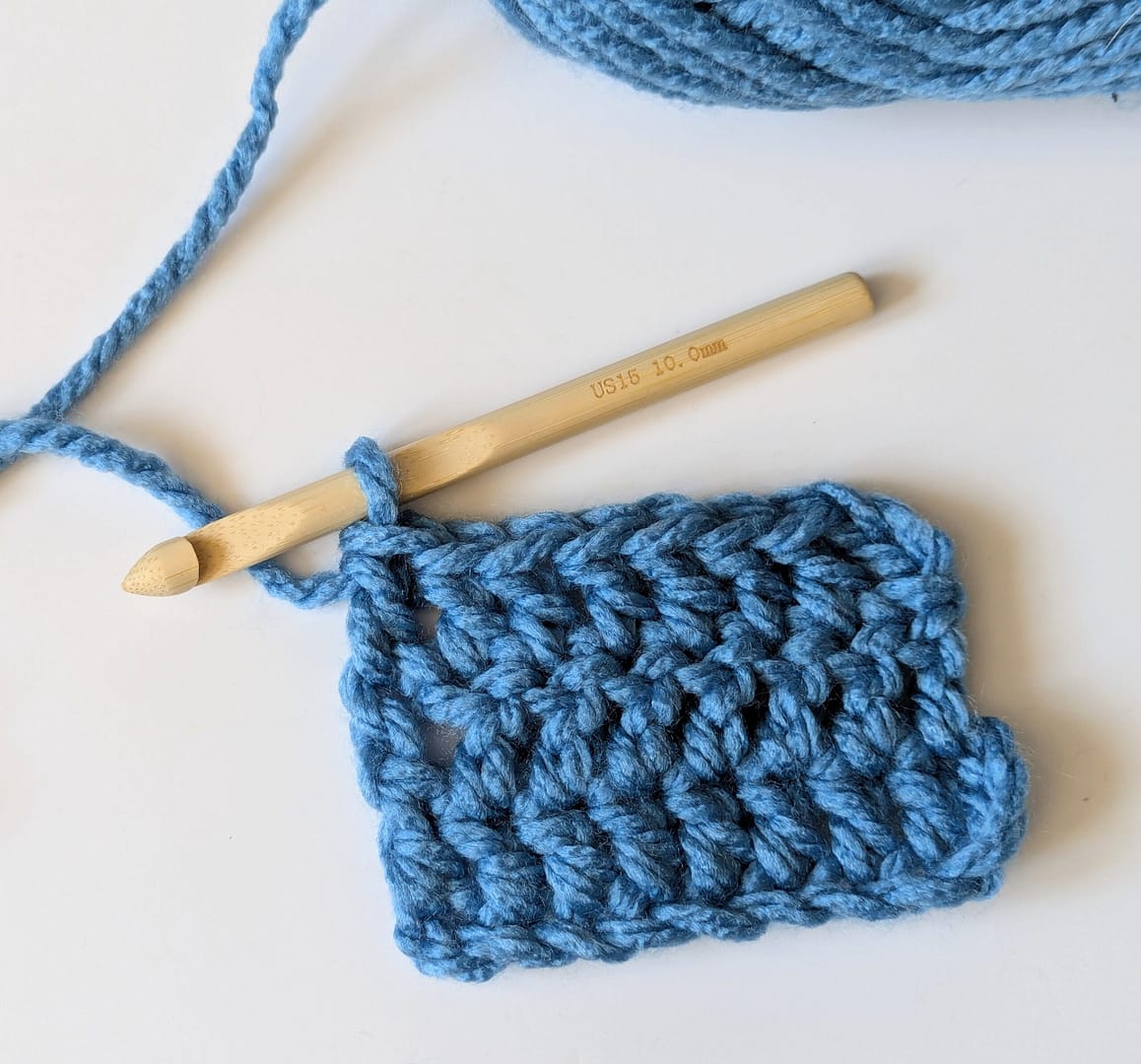 A square of crocheted fabric in blue yarn and a wooden crochet hook is attached