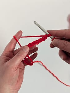 Two hands holding a crochet hook and a run of 15 chain stitches made with red yarn