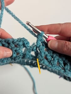 Close of crochet V stitch in blue yarn on a white background, with a yellow stitch marker