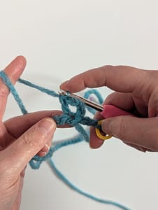 Two hands holding a crochet hook and blue yarn with a stitch in a triangle shape