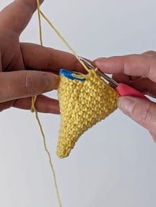 Two hand holding a crochet hook and some single crochet stitches creating the shape of a cone in yellow yarn, there is a blue stitch marker in the first stitch
