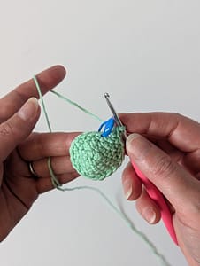 Two hands holding a crochet hook and a small cone shape crocheted fabric in Mint Green yarn