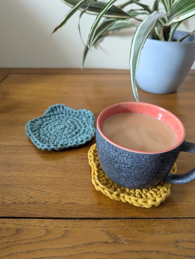 Crochet Coasters, one with a mug of tea on top, on a wooden coffee table and a plant in the background
